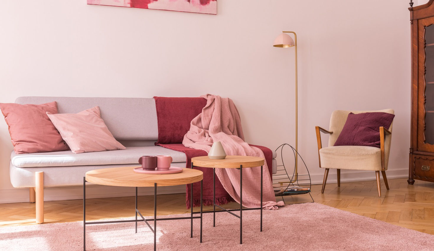 Two wooden coffee tables next to modern grey sofa with pillows and blankets in lovely pastel pink living room interior